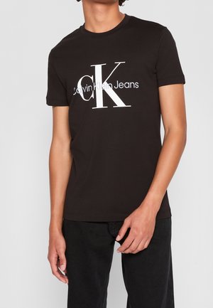 Osoba nosząca czarną koszulkę z krótkim rękawem z dużym białym logo „cK Calvin Klein Jeans”, w połączeniu z czarnymi spodniami na szarym tle.