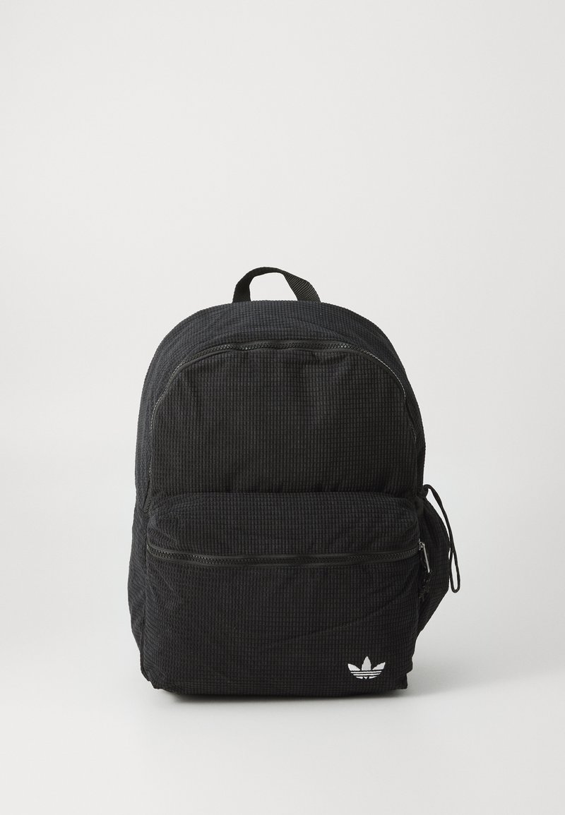 Mochila preta texturizada com formato arredondado, apresentando um bolso frontal com zíper e um bolso lateral. Inclui uma alça superior e detalhe do logótipo.