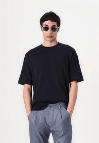 Námořnické tričko s kulatým výstřihem, spojené se šedými upravenými kalhotami s knoflíkem a sklady; model má na sobě sluneční brýle.