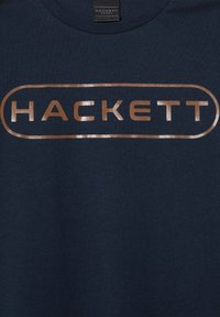 Námornícky modrá košeľa s kovovo bronzovým logom "HACKETT" sústredeným v oválnom obryse a štítkom Hackett Sport na golieri.