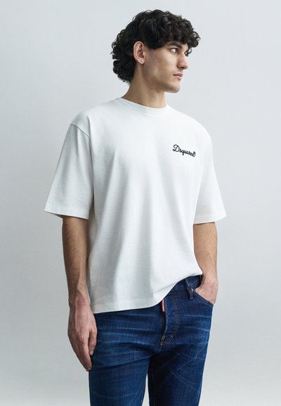 Λευκό βαμβακερό t-shirt με κοντά μανίκια, με μαύρο κεντητό κείμενο στην αριστερή πλευρά· φορεμένο με μπλε τζιν παντελόνι.