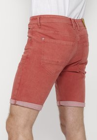Pantalones cortos de mezclilla roja con puños doblados, que cuentan con dos bolsillos traseros y detalles de costura en contraste. La tela tiene una textura desgastada.