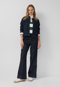 Dunkelblauje Jeansjacke mit Knopfverschluss, zwei Fronttaschen, über einem gestreiften Hemd getragen. Ausgestellte dunkelblaue Jeans, kombiniert mit weißen Turnschuhen.