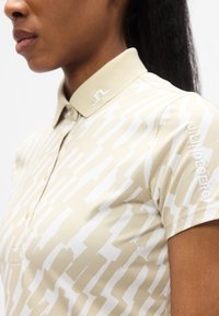 Μπεζ πόλο πουκάμισο με λευκό ζιγκ-ζαγκ μοτίβο, κοντά μανίκια, γιακά με κεντημένο λογότυπο και όνομα μάρκας στο μανίκι.