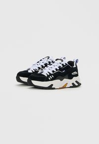 Pair of black and white chunky sneakers with thick soles, white laces, and blue pull tabs, shown on a plain white background.
