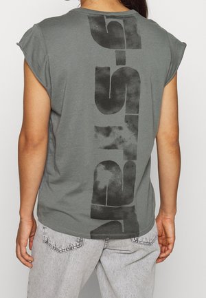 Persona con una camiseta sin mangas gris con letras abstractas verticales negras en la espalda, combinada con jeans gris claro.