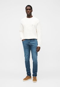 Lichtgekleurde gebreide trui, blauwe spijkerbroek en beige sneakers. Slim fit, gladde textuur, met een klein logo op de trui. Eenvoudig ontwerp.