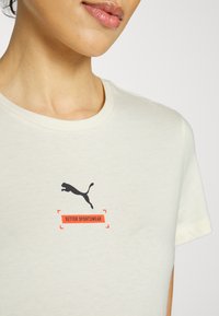 Světle béžové tričko s černým logem Puma nad textem "BETTER SPORTSWEAR" v oranžové barvě. Hladká textura, kulatý výstřih.