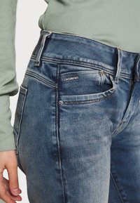 Džínové kalhoty ve středním modrém odstínu s vybledlými akcenty, s klasickým designem pěti kapes a značkovým logem G-Star na přední kapse.