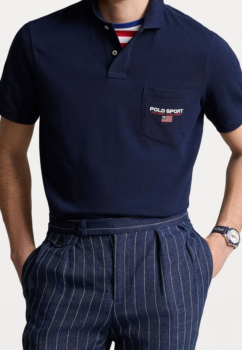 Homme portant un polo bleu marine Polo Sport avec poche poitrine et pantalon rayé bleu marine, mains dans les poches et montre-bracelet au poignet gauche.