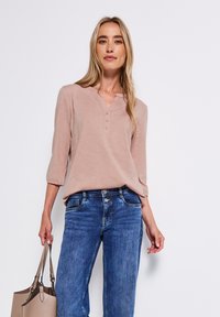 Haut henley à manches longues rose clair avec un col en V et trois boutons, associé à un jean bleu fitted et tenant un sac fourre-tout beige.