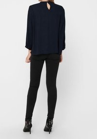 Blusa blu Navy con maniche lunghe, caratterizzata da un dettaglio a chiave sul retro. Abbinata a jeans skinny neri e scarpe con tacco alto. Tessuto dalla texture liscia.
