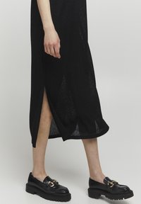 Abito nero lavorato a maglia con spacchi laterali, abbinato a mocassini neri robusti con dettagli in metallo dorato. Tessuto testurizzato, lunghezza alla caviglia.