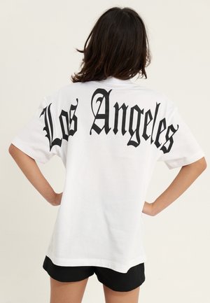 Osoba z ciemnymi włosami zwrócona tyłem, ubrana w białą koszulkę z dużym czarnym napisem "Los Angeles" w stylu Old English na plecach oraz czarne szorty.