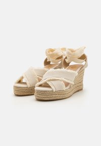Sandalias de cuña con suela de fibra natural, empeine de tela beige, detalles deshilachados y largas cintas de tela para soporte en el tobillo. Superficies texturizadas presentes.