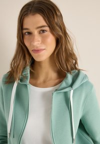 Jeune femme aux cheveux châtain clair ondulés portant un sweat à capuche zippé vert clair sur un haut blanc, fond beige neutre.