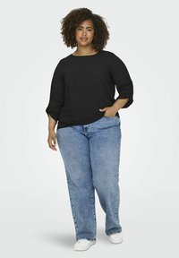 Maglietta nera a maniche lunghe con polsini arrotolati abbinata a jeans azzurri chiari e larghi e sneakers bianche, il tutto su uno sfondo neutro.