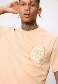 T-shirt en coton pêche à manches courtes avec un graphique floral circulaire vert et le texte "Fleurs & Fragrance" sur la zone gauche de la poitrine.