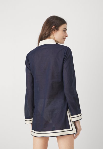 Marinblå tunika med långa ärmar, vit krage och kant. Tillverkad av lätt tyg med en något genomskinlig textur.