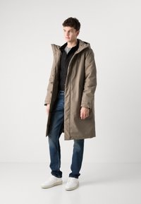 Lang, beige parkajakke med hette, glidelås og sidelommer, kombinert med mørke jeans og hvite joggesko. Glatt tekstur og enkelt design.