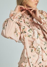 Blusa floreale in morbido tessuto rosa presenta dettagli con volant, maniche lunghe a sbuffo e bottoni decorativi. Le foglie verdi completano il motivo floreale.