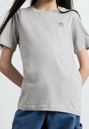 Kobieta ubrana w jasnoszarą koszulkę Adidas z logo na piersi, w połączeniu z niebieskimi jeansami, stoi z rękami za plecami.