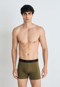 Herren-Boxershorts aus olivgrünem Stoff mit einem schwarzen Bund, der das rote "HUGO"-Logo zeigt. Glatte Textur, figurbetontes Design und mittlere Oberschenkellänge.