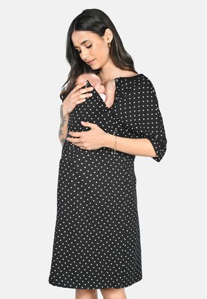 Frau in schwarzem Kleid mit weißen Herzen hält Baby nah an der Brust und wiegt das Kind schützend und sanft auf einfachem Hintergrund.