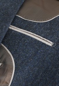 Wollstoff in Dunkelblau mit hellblauen Flecken. Hat eine glatte Textur, mit einem braunen Paneel-Akzent und hellen Nähdetails.