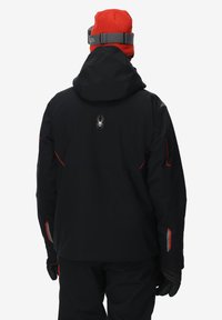 Zwarte waterdichte jas met een capuchon, rode accenten en een spinlogo op de achterkant. Gecombineerd met een rode gebreide muts en zwarte handschoenen.