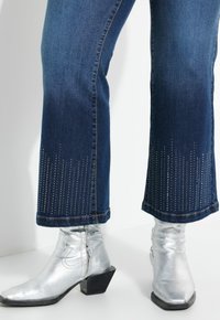 Vaqueros acampanados de mezclilla azul oscuro con acentos de strass en el dobladillo, combinados con botines de tobillo plateados brillantes que cuentan con un tacón cuadrado.