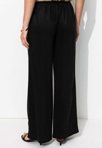 Pantalons noirs à jambes larges en tissu lisse et léger. Taille élastique avec peu de détails. Portés avec des talons aiguilles noirs.