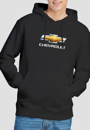 Junger Mann, der einen schwarzen Kapuzenpullover mit einem großen Chevrolet-Logo und dem Wort "CHEVROLET" in Weiß trägt, kombiniert mit blauen Jeans.