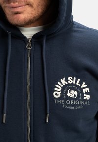 Tmavě modrá mikina na zip vyrobená z měkké tkaniny, se vzorem loga a šňůrkami s kovovými konci. Hladká textura a ležérní design.