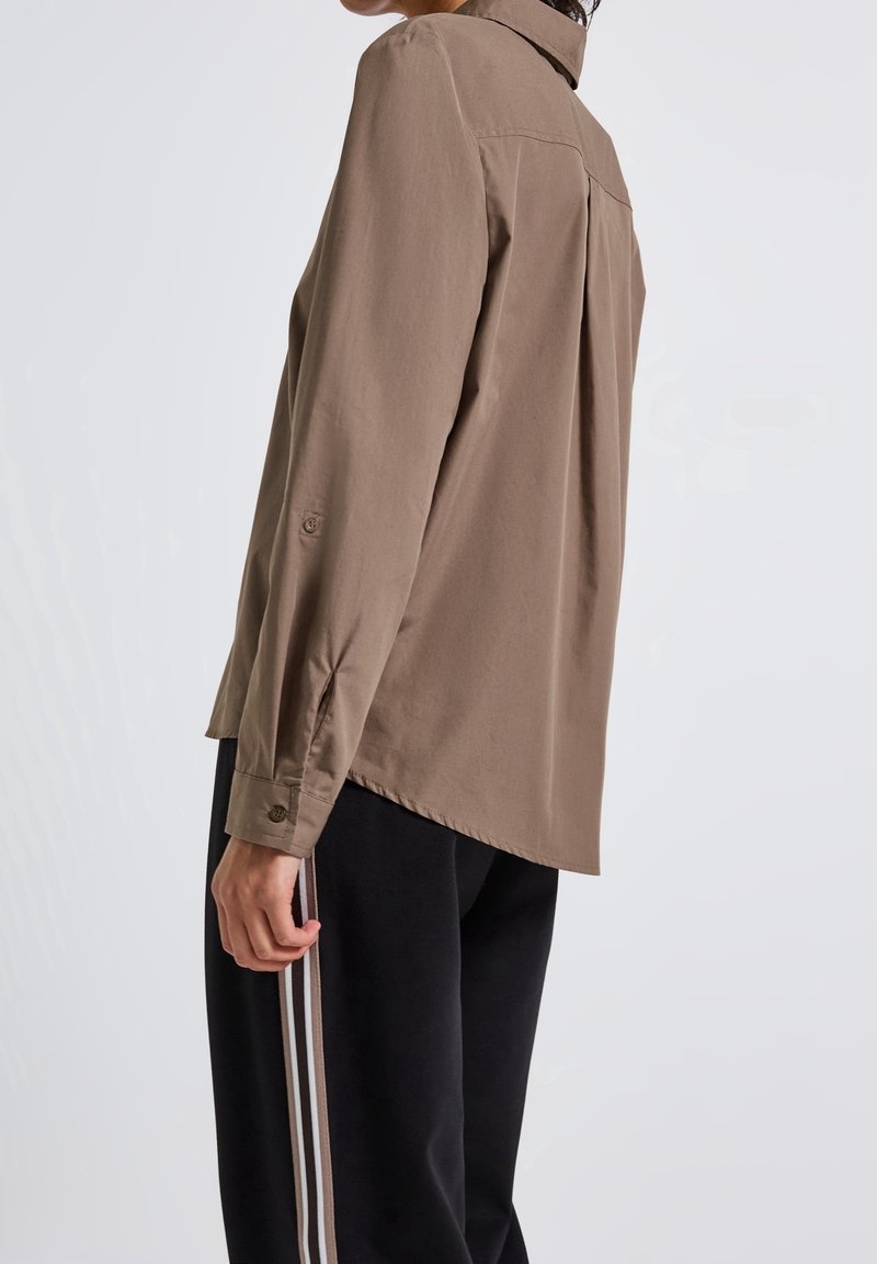 Personne portant une chemise marron ample boutonnée et un pantalon noir avec des bandes latérales blanches et marron, debout de côté devant un fond uni.