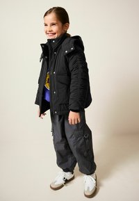 Schwarze Steppjacke mit Reiß- und Druckknopfverschluss, zwei Fronttaschen, kombiniert mit grauen Cargo-Hosen und weißen Sneakers, vor einem neutralen Hintergrund.