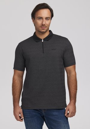 Mężczyzna ubrany w ciemnoszare polo z krótkim rękawem, czarnym kołnierzem i półzamkiem, w połączeniu z niebieskimi dżinsami, stojący na jasnym tle.
