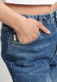 Pantalones vaqueros de mezclilla azul con un diseño ajustado, costuras visibles y un bolsillo frontal. El material parece duradero con una ligera textura.