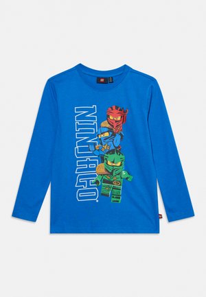 Chemise bleue à manches longues avec des personnages LEGO Ninjago en rouge, bleu et vert, et le mot NINJAGO imprimé verticalement sur le devant.