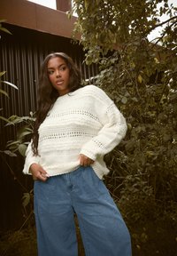 Kremowy sweter dzianinowy z teksturowymi paskami, w połączeniu z luźnymi niebieskimi spodniami dżinsowymi. W tle znajdują się zielone liście oraz czarna pionowa powierzchnia.