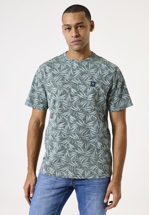 Camiseta verde con un patrón de hojas, de manga corta, con cuello redondo y un parche de tela en el pecho. Lleva puestas unas jeans azules.