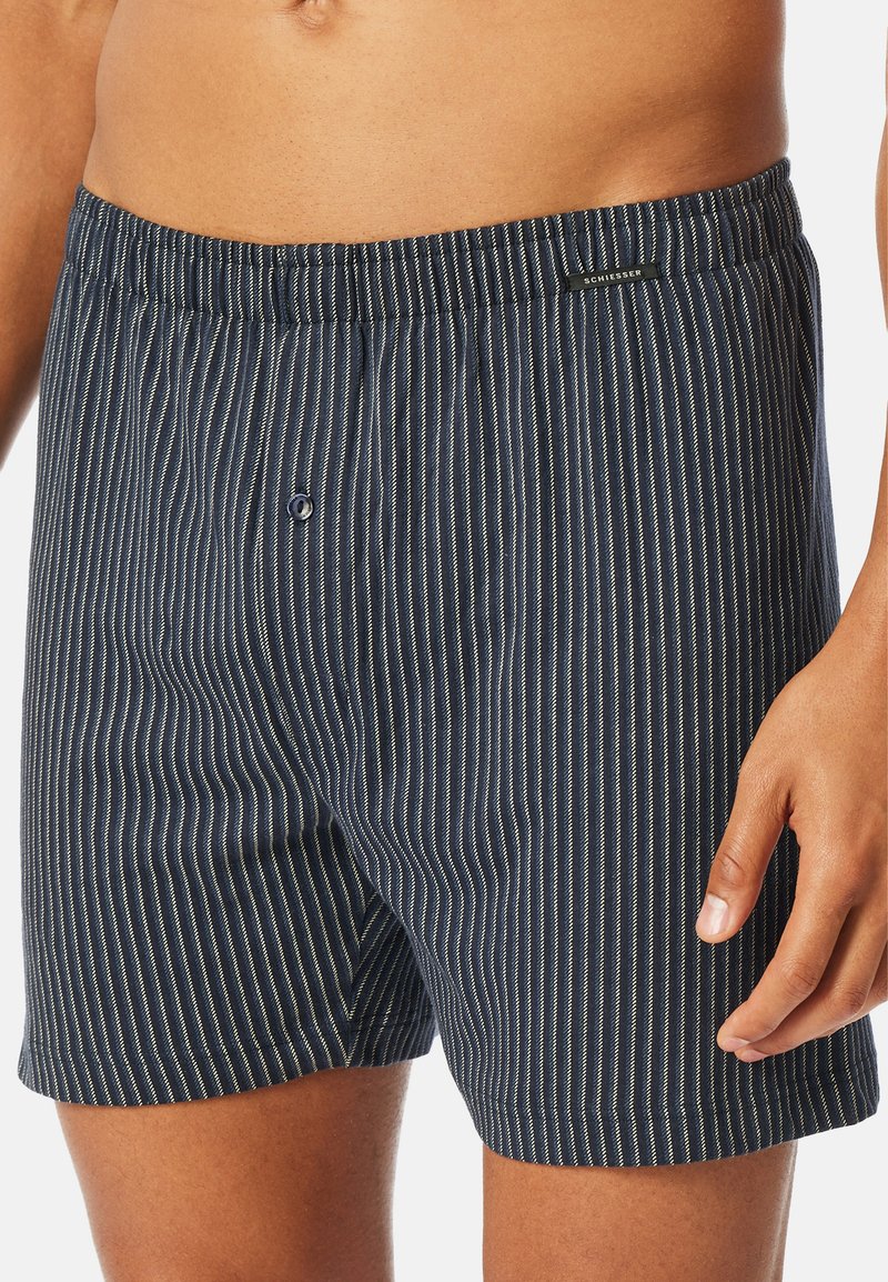 Boxer shorts blu navy con sottili righe verticali bianche, fascia elastica in vita, chiusura con un singolo bottone e materiale in cotone morbido.