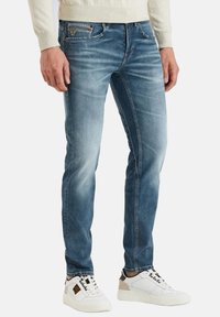 Jeans azules desgastados con un corte slim y detalles sutiles de arrugas, combinados con zapatillas blancas con acentos en negro y beige.