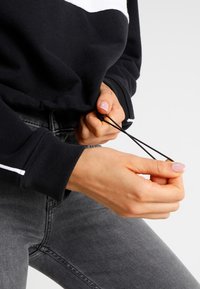 Felpa nera con accenti bianchi, cordino elastico tirato, indossata con jeans grigi. Tessuto morbido e polsini a coste visibili.