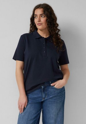 Kobieta o kręconych włosach, ubrana w granatową koszulkę polo i niebieskie dżinsy, stojąca z jedną ręką w kieszeni na jednolitym tle.