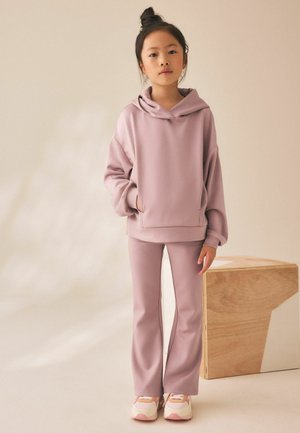 Junges Mädchen mit Dutt, trägt einen hell mauvefarbenen Hoodie und passende Hose, steht neben einem hölzernen geometrischen Hocker vor neutralem Hintergrund.