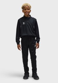 Junge mit blonden Dreadlocks, der einen schwarzen Hummel-Trainingsanzug und schwarze Sneaker trägt, steht lächelnd vor einem schlichten hellen Hintergrund.