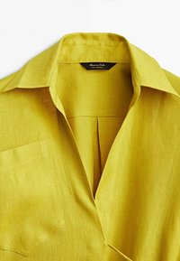 Lyhythihainen, muotoiltu sinapinkeltainen paita, joka on valmistettu pellavasta ja jossa on terävä kaulus, rintatasku ja hienovarainen kuitu.