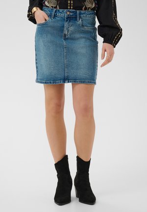 Vrouw draagt een blauwe denim minirok, een zwarte geborduurde blouse en zwarte suède enkellaarzen, staat met één hand in de zak.