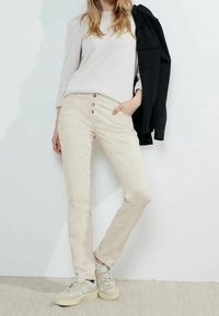 Beżowe bawełniane spodnie o dopasowanym kroju, z pięcioma guzikami z przodu i kieszeniami po bokach, zestawione z jasnoszarym swetrem i beżowymi trampkami.